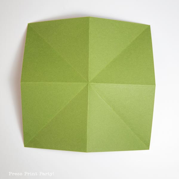 A green piece of paper, expertly folded with intersecting diagonal and vertical lines creating triangular sections, forms a symmetrical pattern reminiscent of a Christmas tree, all placed on a pristine white background.