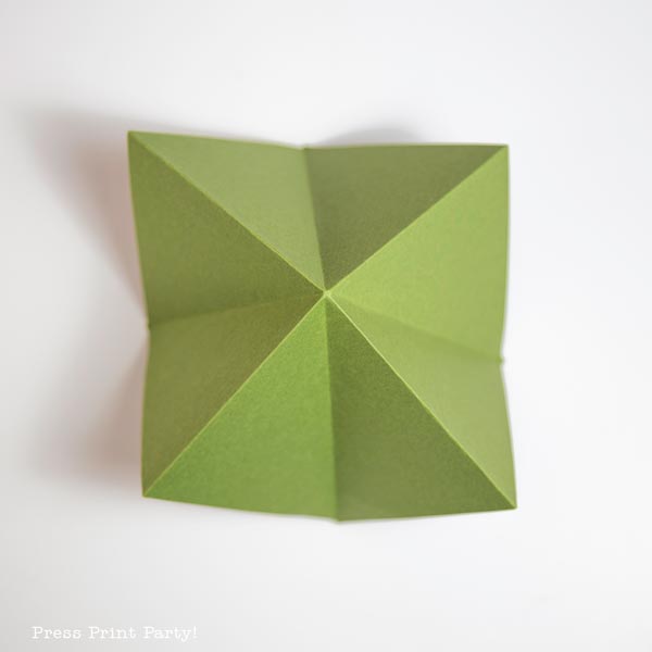 A folded paper Christmas tree, resembling a fortune teller with four triangular peaks, is placed on a white surface.