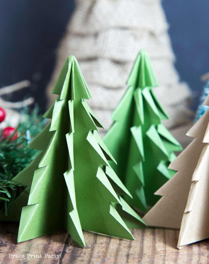 Close-up of folded paper Christmas trees in green and beige on a wooden surface. The intricately layered, pointed branches add charm, while blurred decorations in the background suggest a festive setting.