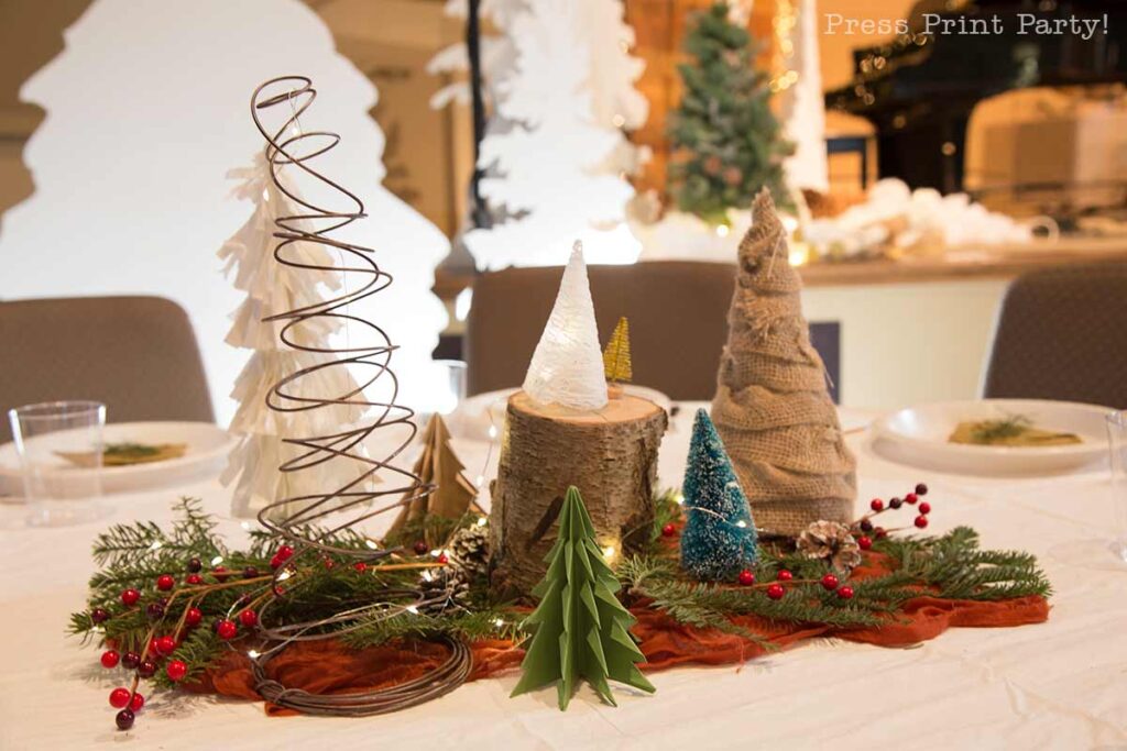 A festive holiday table centerpiece showcases folded paper Christmas trees crafted from wire, fabric, and paper, elegantly arranged on a white cloth. The display is accented with lush green pine branches, vibrant red berries, and the inviting glow of warm string lights.