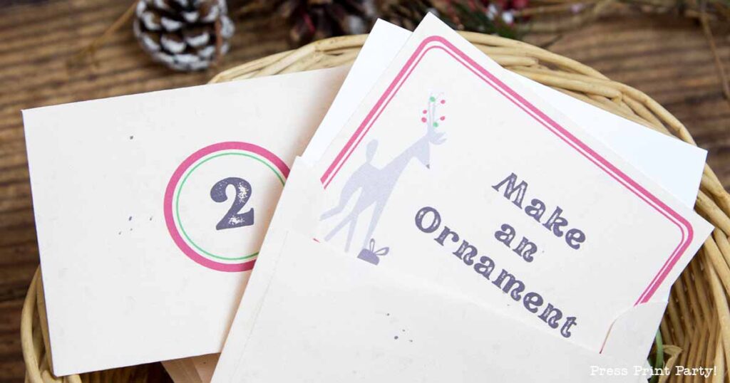 A close-up of a basket holding holiday-themed cards. One card shows the number "2" in a circle, and another card reads "Make an Ornament" with a reindeer illustration. Pinecones and greenery are visible in the background.