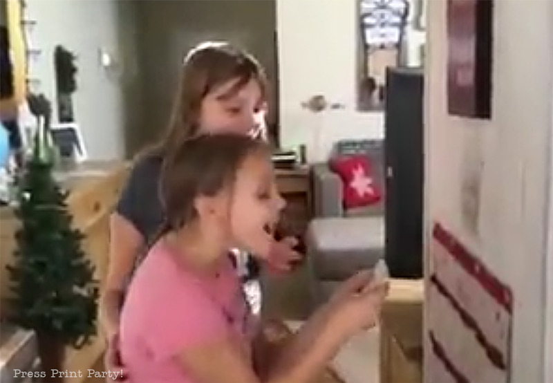 Two young girls excitedly open an advent calendar in a cozy living room. A small decorated Christmas tree is visible in the background, along with a couch and festive décor.