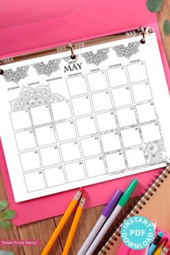 The desk scene showcases a 2025 One Page Calendar at-a-glance Printable Mandala, embellished with intricate designs, enclosed in a pink binder. Adjacent to it are vibrant pens, a spiral notebook, and green leaves. A turquoise sticker announces 