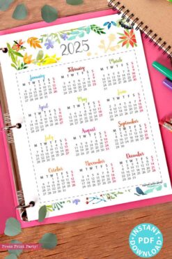 A 2025 calendar printable page, featuring a watercolor design, is displayed on a spiral notebook with colorful floral borders. Each month is laid out in a grid format. The notebook is placed on a pink surface, surrounded by green leaves and vibrant pens. A badge reading 