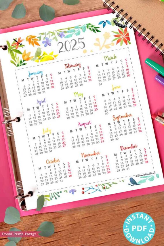 A 2025 calendar printable page, featuring a watercolor design, is displayed on a spiral notebook with colorful floral borders. Each month is laid out in a grid format. The notebook is placed on a pink surface, surrounded by green leaves and vibrant pens. A badge reading "Instant PDF Download" decorates the corner.