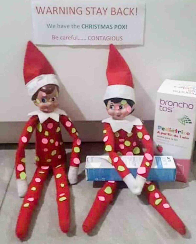 Two toy elves in red outfits with colorful spots sit beside a box labeled "bronchotos." A sign above reads, "WARNING STAY BACK! We have the CHRISTMAS POX! Be careful... CONTAGIOUS.