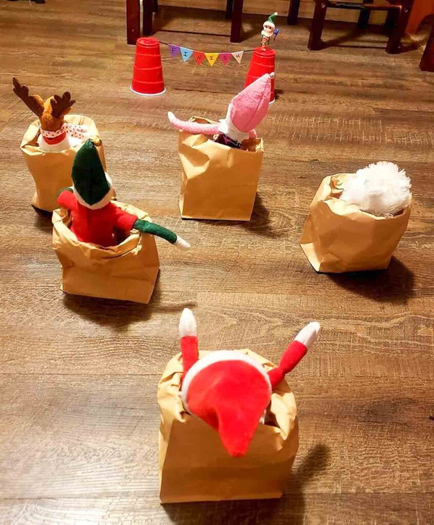 Stuffed toys dressed as elves and animals are playfully sitting in brown paper bags on a wooden floor. A small festive banner and red plastic cups are visible in the background, adding a playful, celebratory atmosphere.