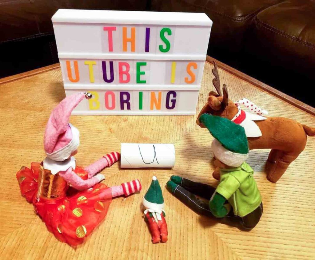 Three plush characters, including a reindeer and two elves, sit around a paper scroll on a wooden table. A lightbox sign reads "THIS YOUTUBE IS BORING" with colorful letters.