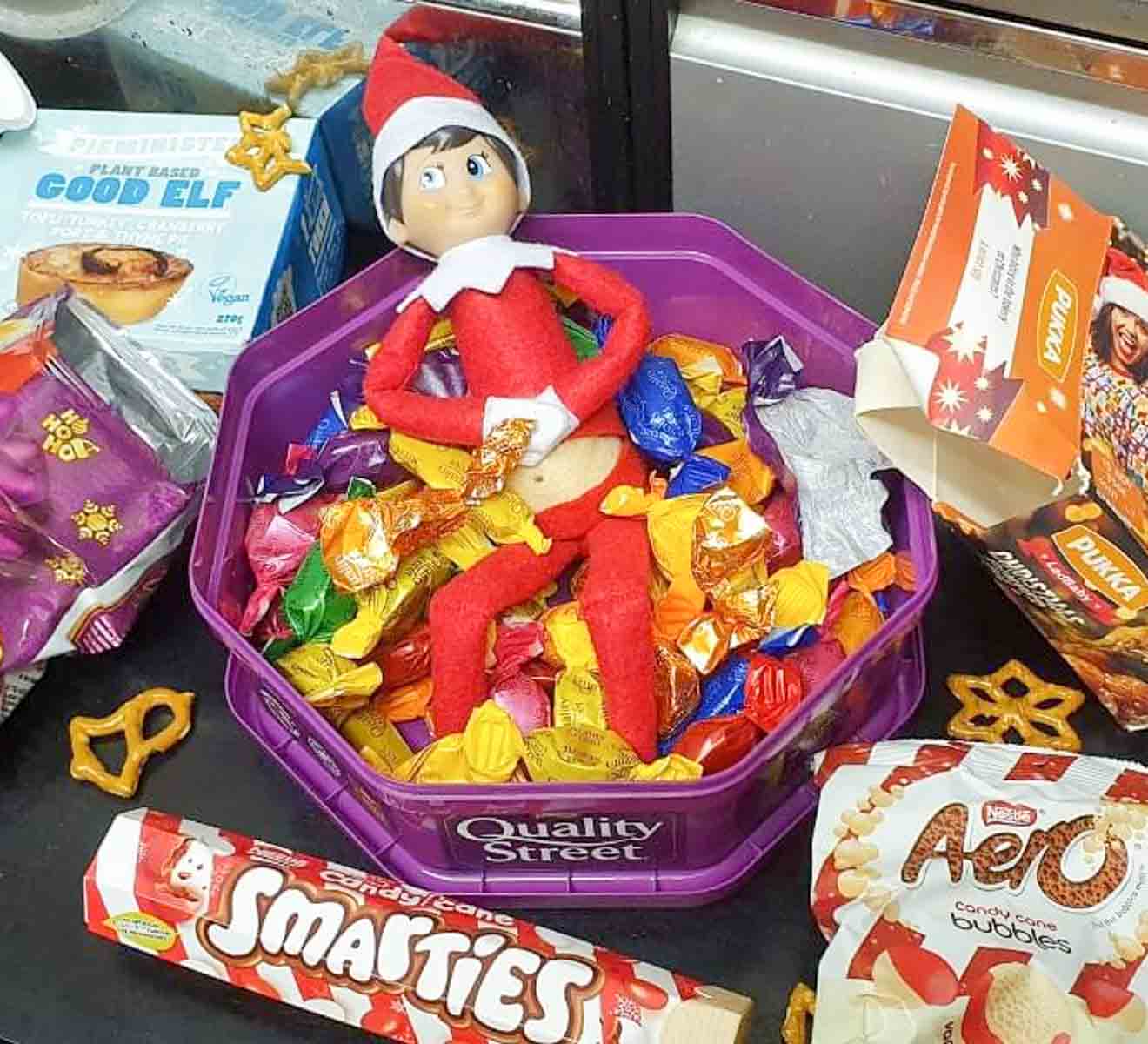 A small elf doll lies on a pile of assorted candies in a purple Quality Street tin. Surrounding the tin are various candy packages, including Smarties and Aero. Some pretzels are scattered around the scene.