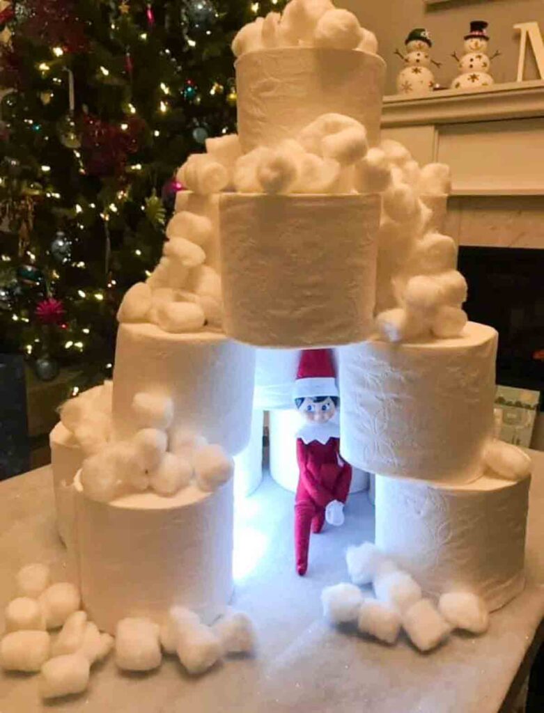A festive scene featuring a toy elf sitting inside a pyramid made of toilet paper rolls. The rolls are topped with cotton balls. A decorated Christmas tree and snowman figurines are in the background.