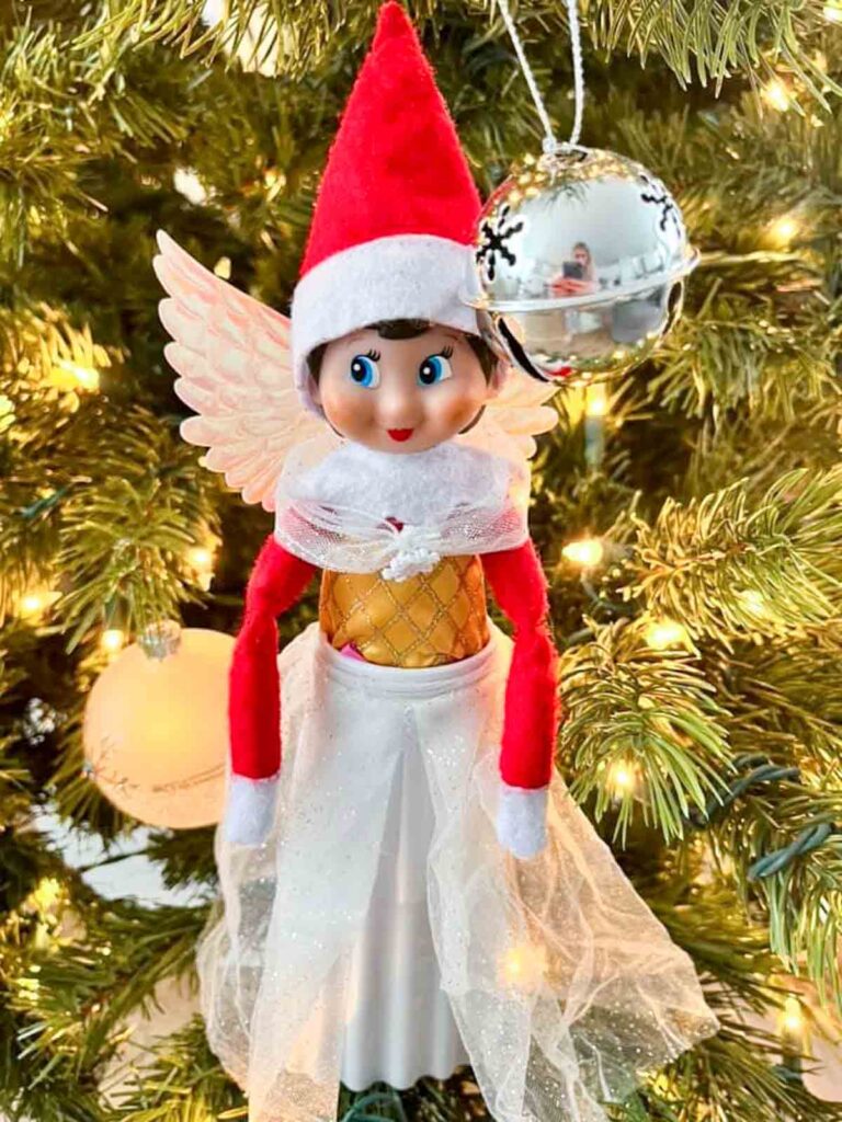 A festive elf doll wearing a Santa hat and angel wings is perched on a Christmas tree. It is dressed in a red and gold outfit with a white skirt, and is surrounded by sparkling lights and ornaments, including a shiny silver bauble.