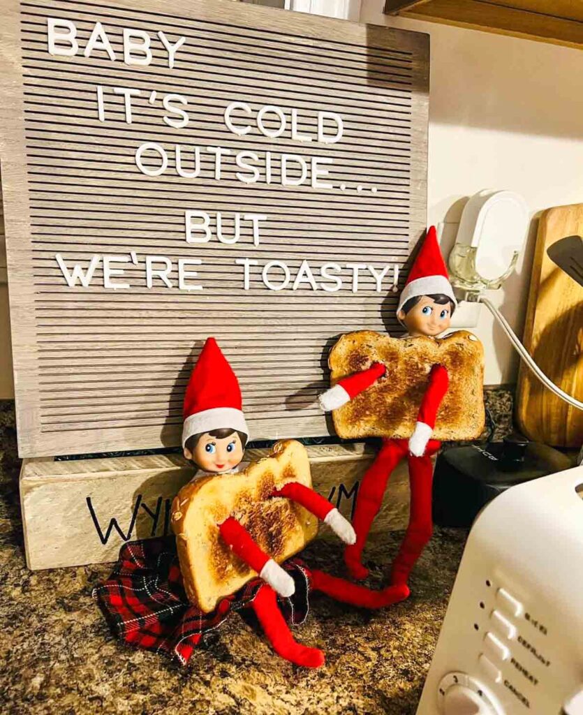 Two elf dolls in red outfits sit on a countertop holding plush toast slices. Behind them is a board with the message "Baby it's cold outside...but we're toasty!" A toaster is visible in the foreground.