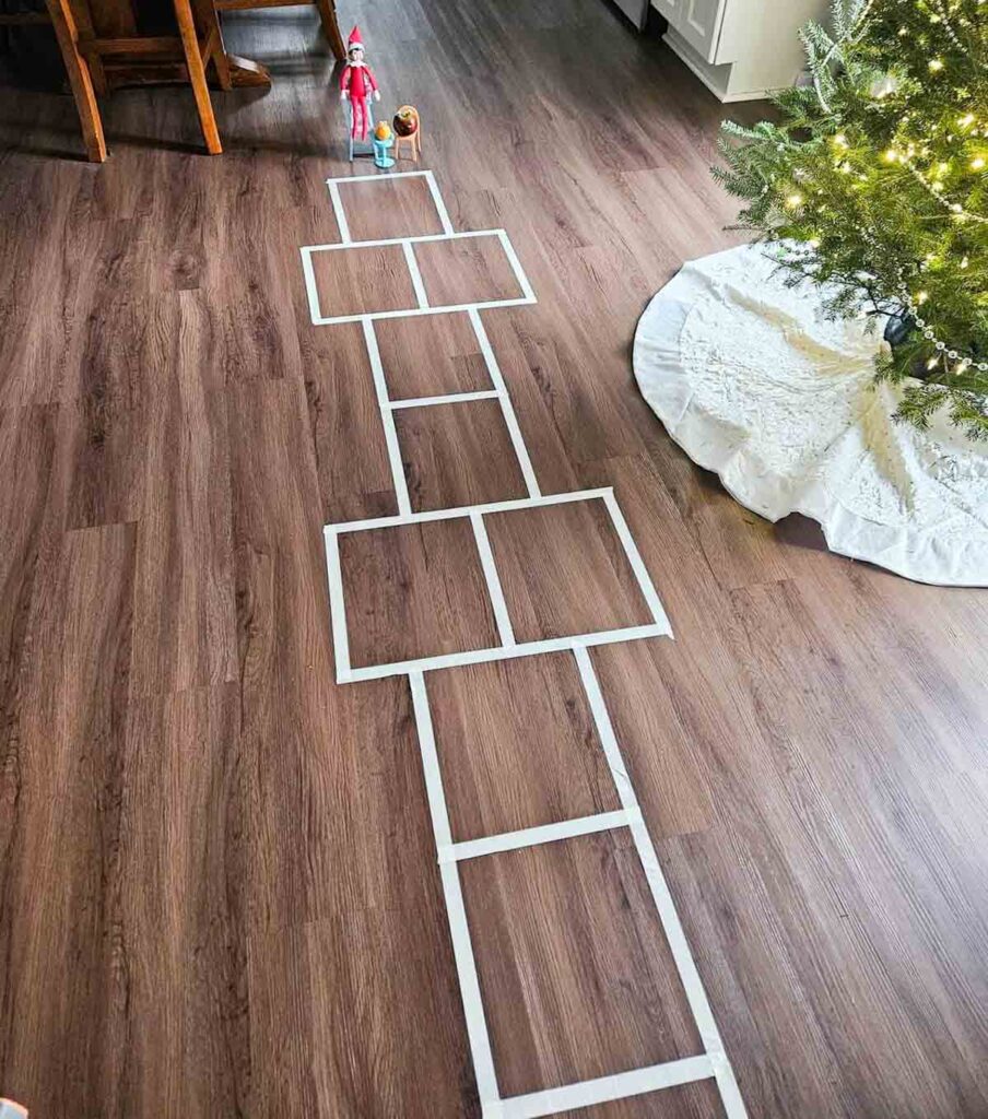 A hopscotch pattern made of white tape is laid out on a wooden floor. A small doll and an elf figure stand at the start. A Christmas tree with white tree skirt is visible on the right.