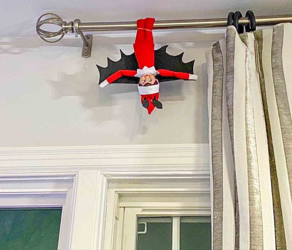 A small elf doll dressed as a bat hangs upside down from a curtain rod. A striped curtain is partially visible beside it.