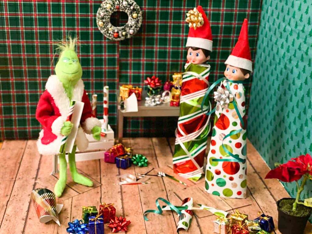 A festive scene with a green character in a Santa outfit sitting on a chair, surrounded by Christmas decorations and gifts. Two dolls dressed as elves are wrapped in colorful gift paper, standing near a small table with more decorations.