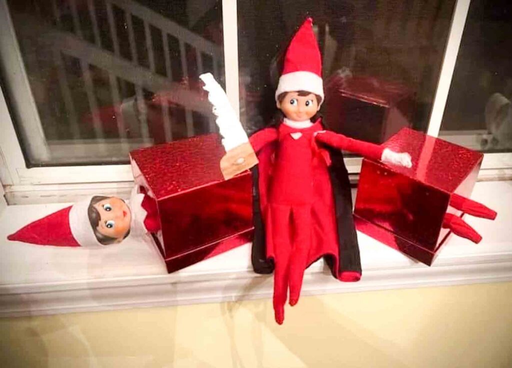 A mischievous elf figurine dressed in red sits on a windowsill holding a small paper knife, surrounded by two red gift boxes. The boxes have elf heads popping out, creating a playful and whimsical scene.