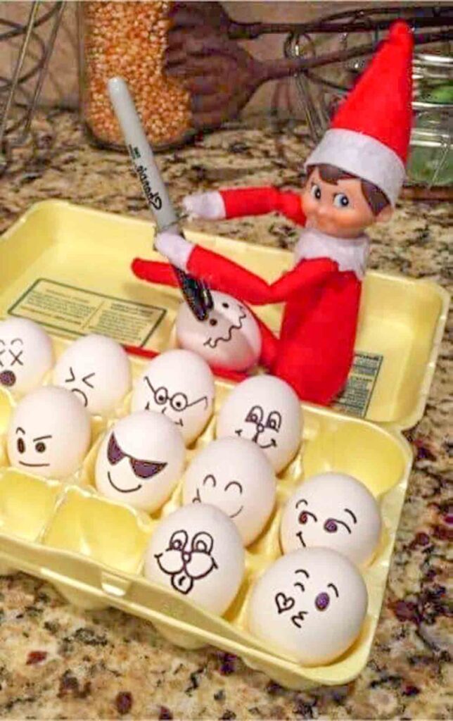 A mischievous elf toy is drawing faces on a dozen white eggs in a yellow carton. The elf is holding a black marker, and each egg has a different playful expression. The scene is set on a kitchen counter.