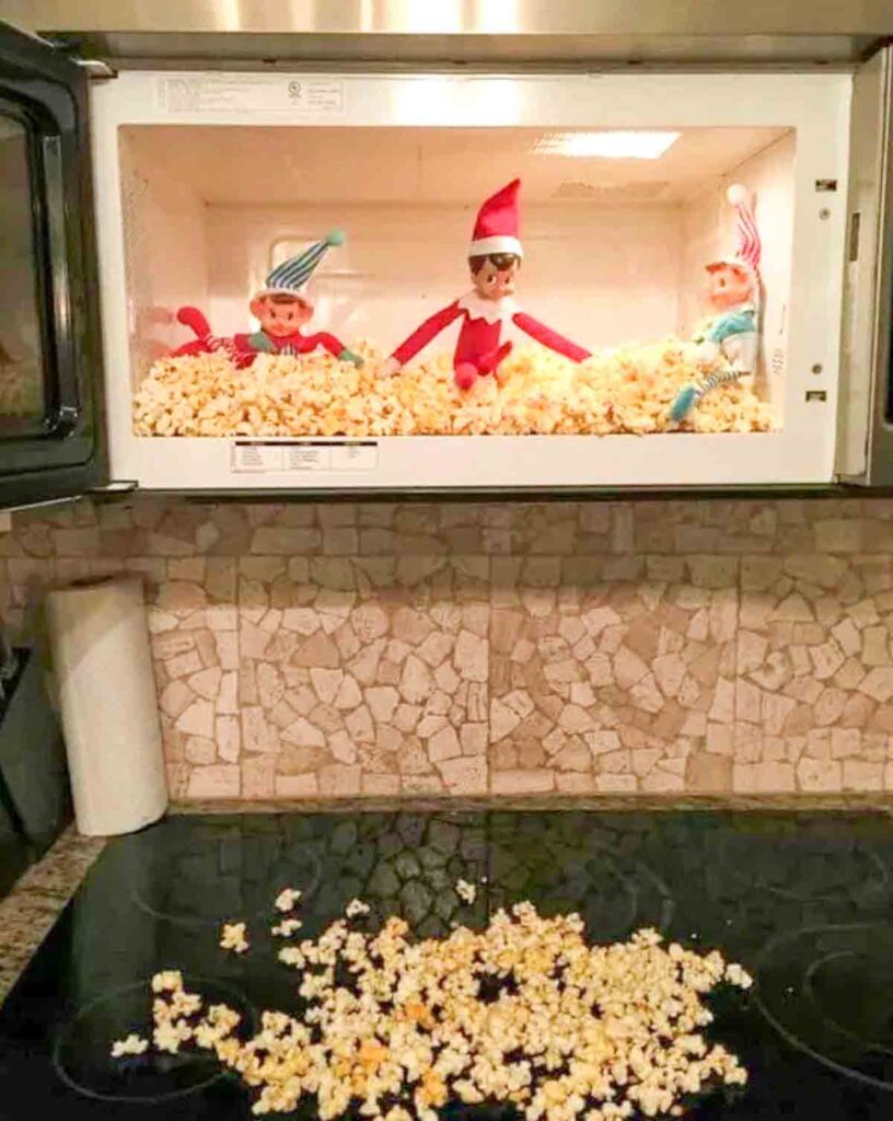 Three toy elves sit inside a microwave filled with popcorn, with some popcorn spilling onto the stove below. The elves are wearing festive hats and appear to be in a playful scene.