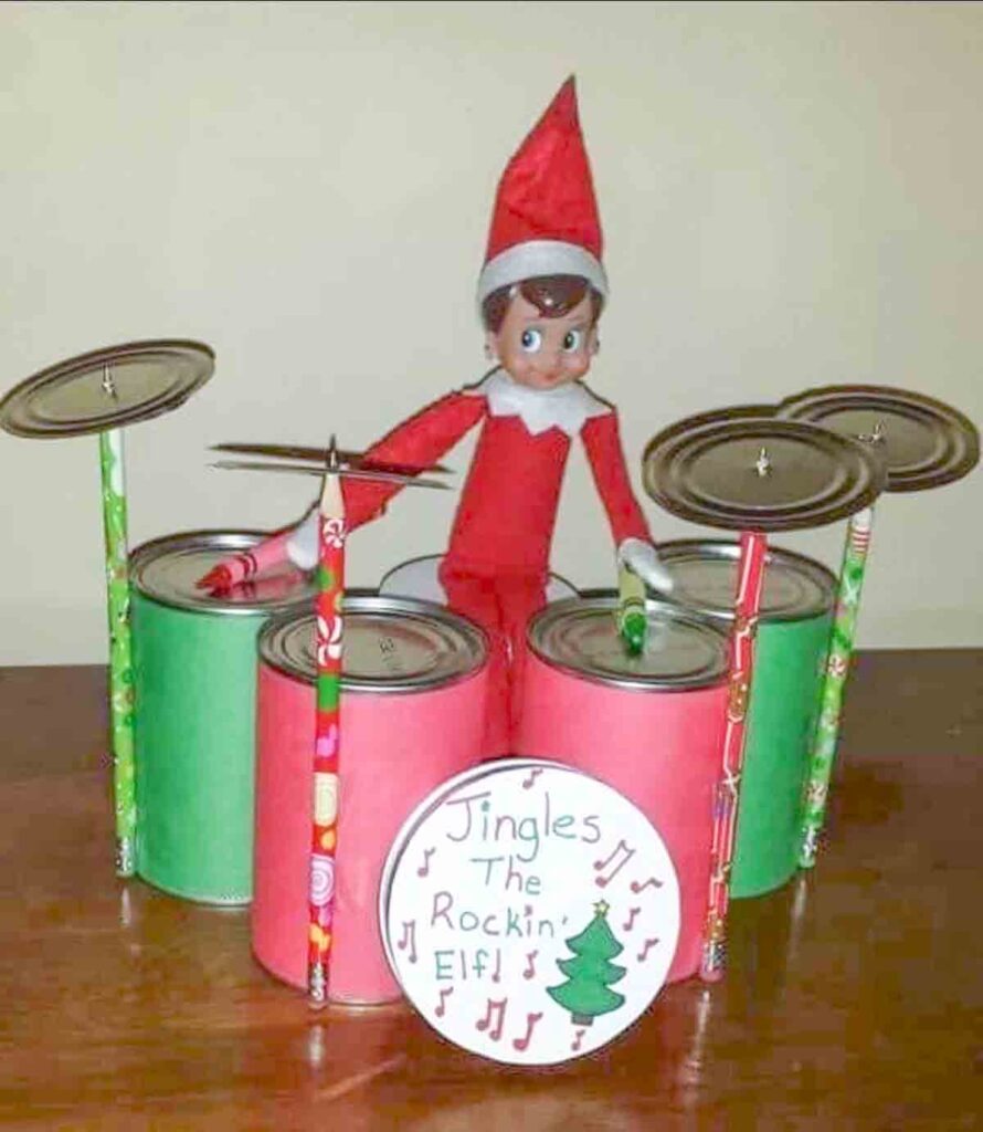 An elf doll is sitting on a makeshift drum set made of colorful cans and pencils. The drum set has a sign saying "Jingles the Rockin' Elf!" with musical notes and a Christmas tree drawn on it.