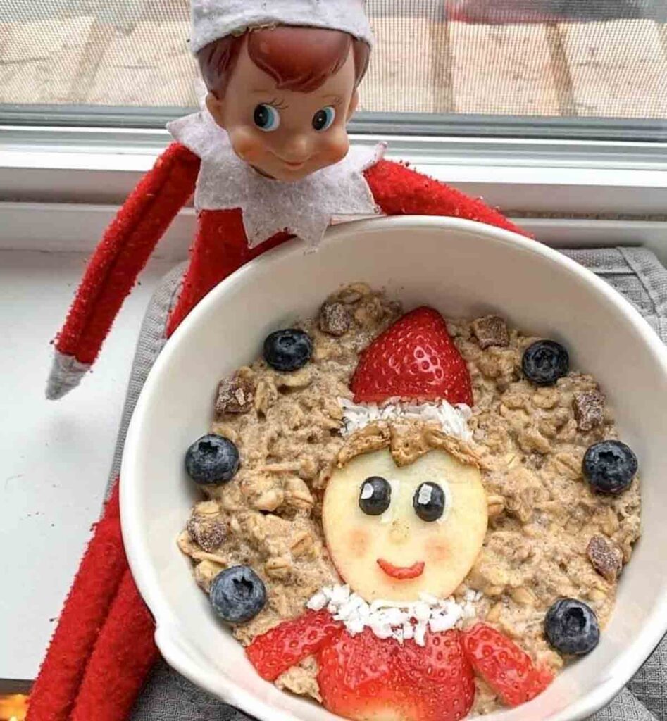 A festive bowl of oatmeal with a face design made from fruit. Strawberry and apple slices form the hat and face, blueberries as eyes, and yogurt for the smile. An elf doll sits nearby on a windowsill.