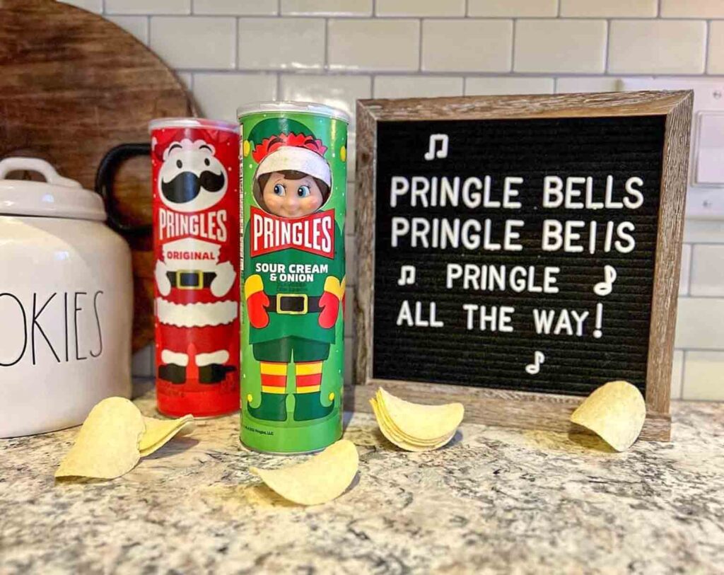 Two festive Pringles cans, one with Santa and another as an elf, are on a counter with several chips. A letter board reads, "Pringle Bells Pringle Bells Pringle all the way!" A cookie jar is nearby against a tiled backsplash.