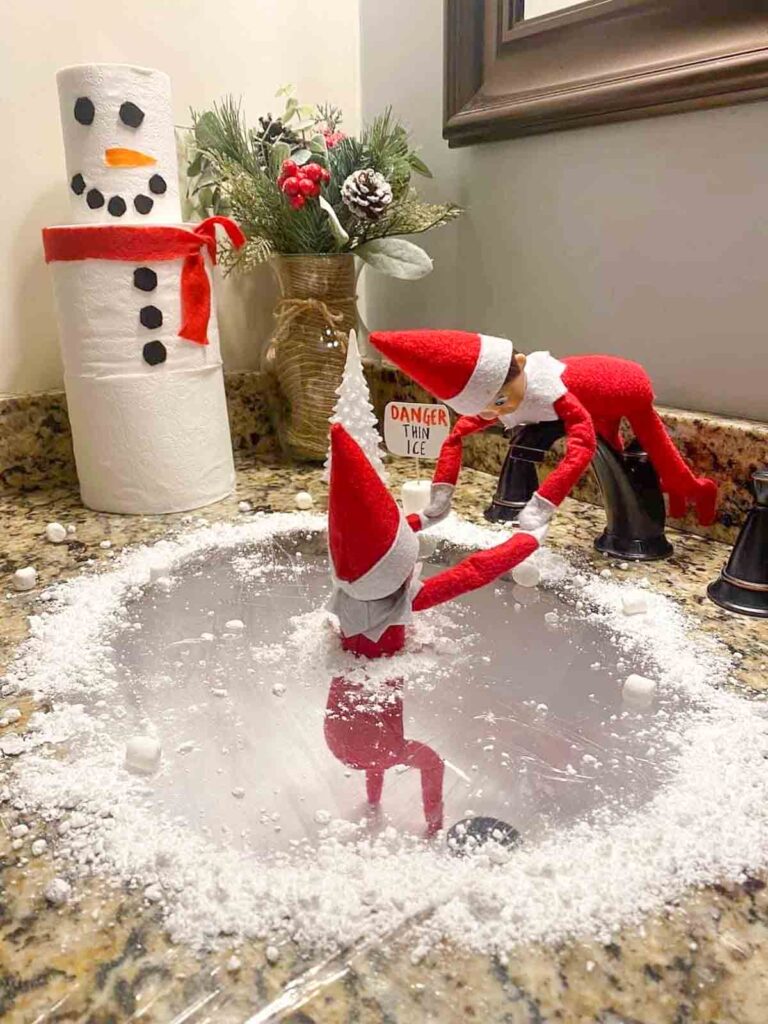 Two elf dolls are playfully posed on a bathroom sink covered with plastic wrap and fake snow, mimicking swimming in a frozen pond. A decorated paper towel roll resembling a snowman is in the background, adding a festive touch.