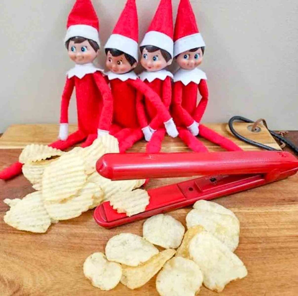 Four elf dolls in red outfits sit on a wooden surface behind a pile of potato chips. A red hair straightener lies in front, suggesting it's been used humorously to make the chips.