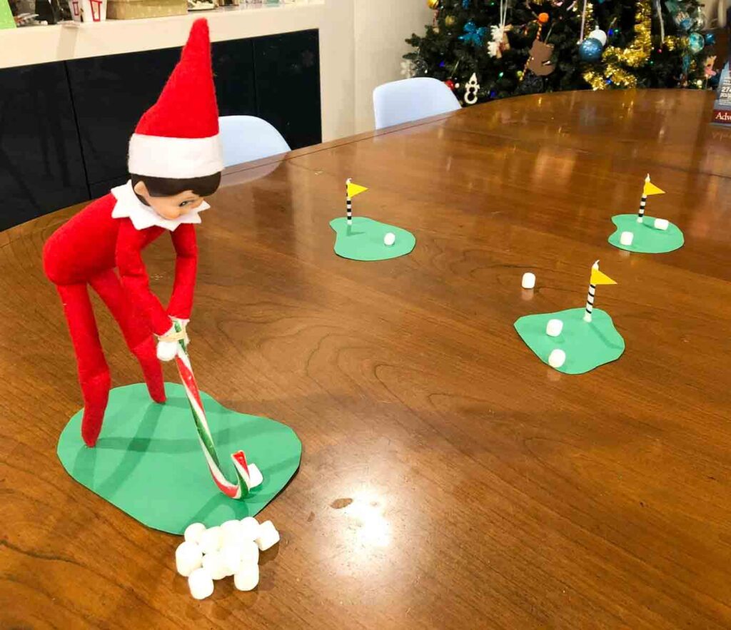 A toy elf dressed in red stands on a wooden table, using a candy cane like a golf club to play mini-golf with marshmallows. Small green paper "greens" with white balls and flagsticks are set up as holes. A Christmas tree is in the background.