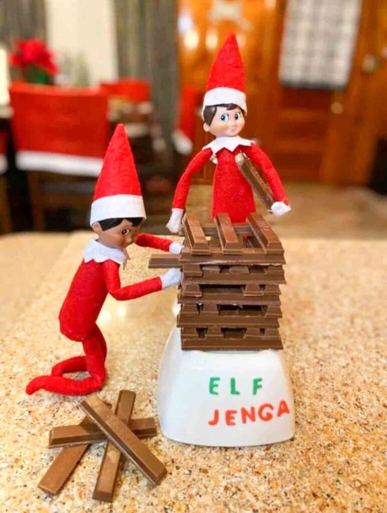 Two elf dolls dressed in red and white play a game resembling Jenga with stacked wooden sticks on a white base labeled "Elf Jenga." One elf is kneeling, and the other is standing next to the stack.