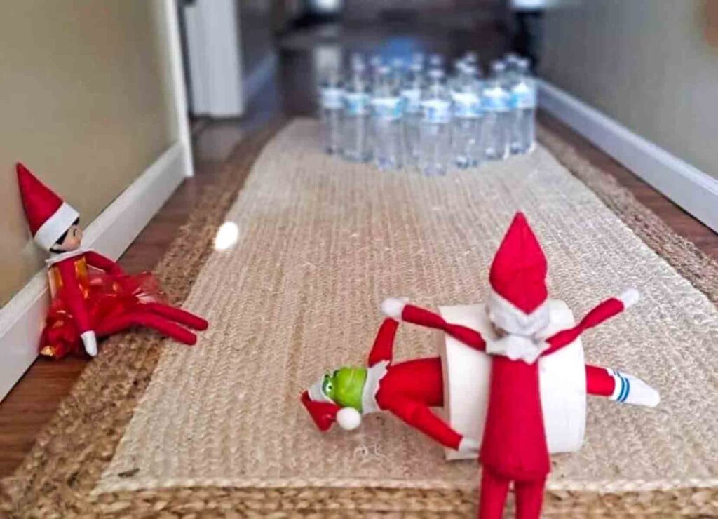 Two elf dolls are on a rug: one is sitting, and the other, wrapped in toilet paper, is lying down. They face a set of water bottle pins at the end of the rug, resembling a bowling game.