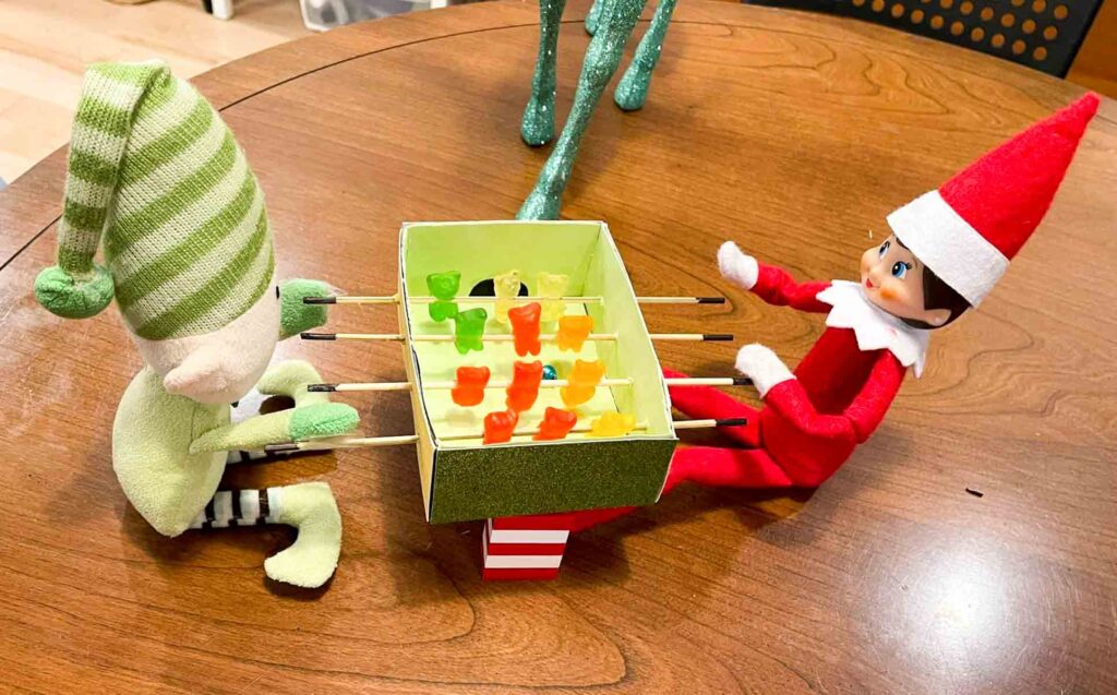 Two elves are playing foosball with gummy bears on skewers in a makeshift table. One elf is dressed in green and white, the other in red. They are sitting on a wooden table, and there is a chair and green decor in the background.