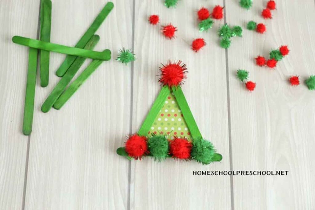A delightful DIY Christmas craft, this green popsicle stick triangle adorned with red and green pom-poms resembles a festive tree. It's perfect as an "Elf on the Shelf" craft, with extra sticks and pom-poms scattered on a light wood background for added creative fun.