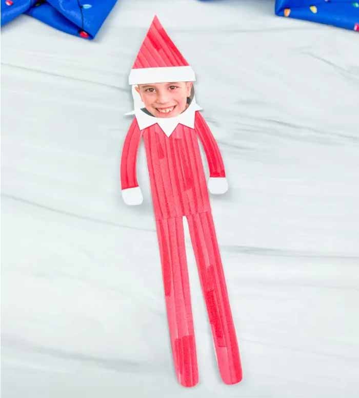 Craft stick transformed into a delightful Elf on the Shelf creation, showcasing a smiling child's face adorned with a red and white costume and pointed hat, all part of enchanting elf on the shelf crafts. It rests charmingly on a light surface, ready to spread holiday cheer.