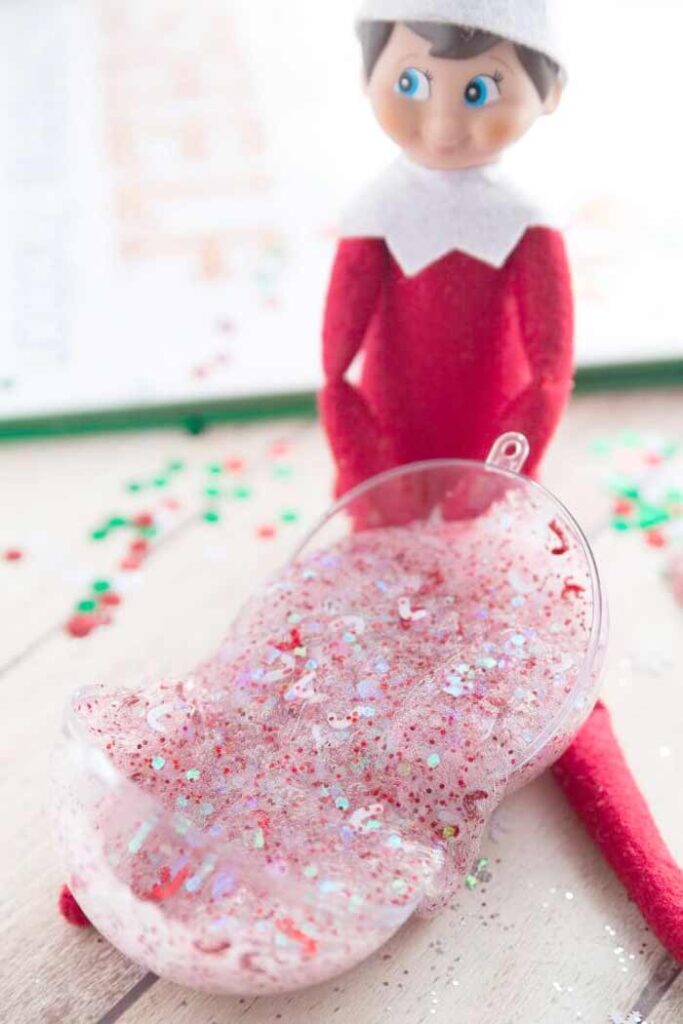 A red-suited elf doll, perfect for "Elf on the Shelf" crafts, sits next to a clear ornament filled with red and white glittery slime. The festive background is sprinkled with vibrant red, green, and white confetti.