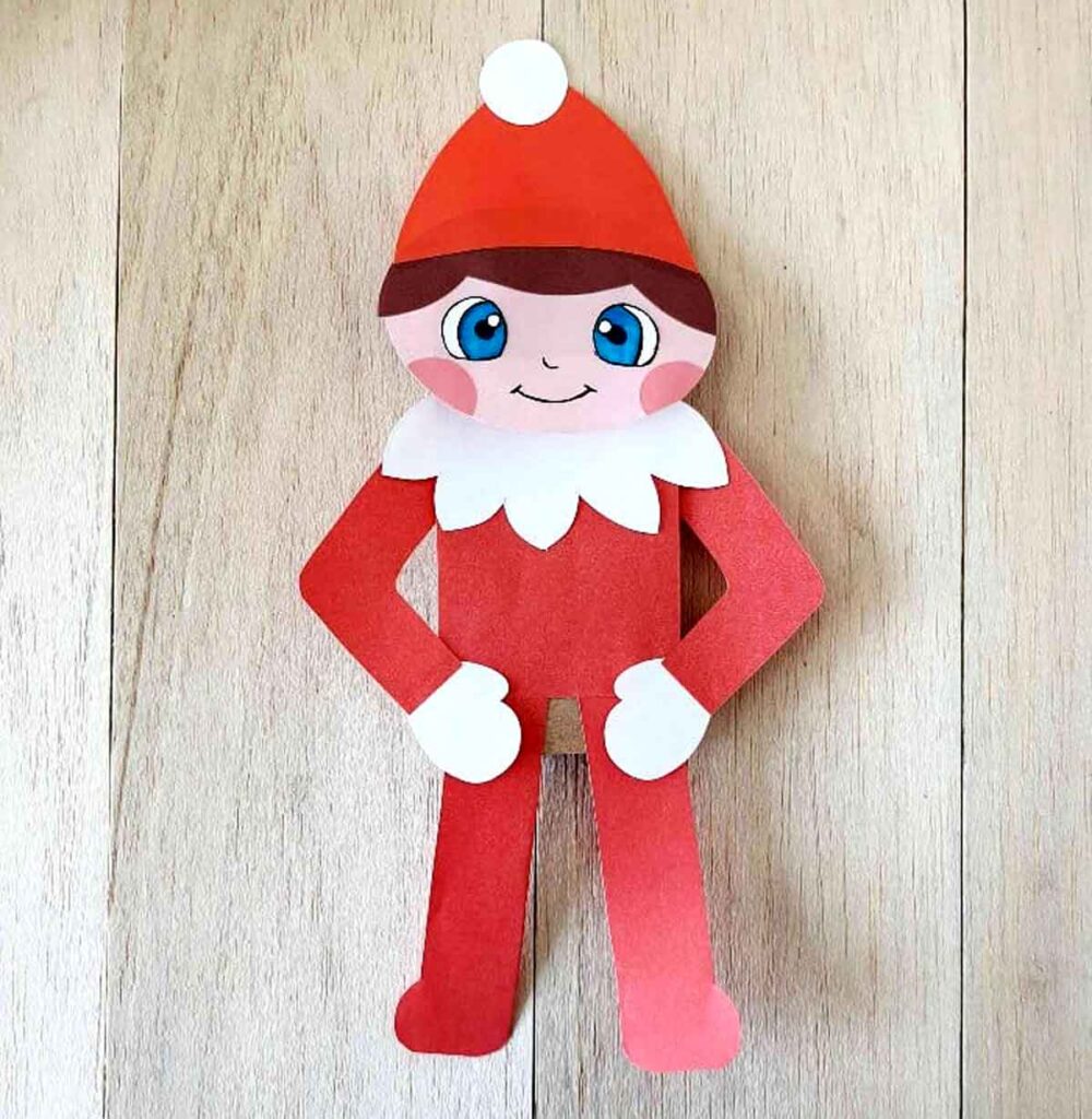 A paper cutout of an elf on the shelf, adorned with a red hat and outfit, white collar, and mittens. Its pink cheeks and blue eyes bring a whimsical charm to this delightful craft piece, standing proudly on a wooden surface.