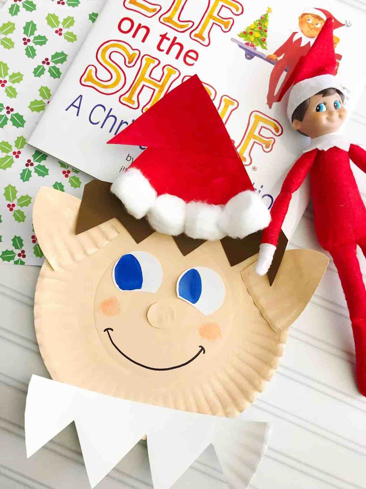 A delightful paper plate craft resembling an elf with a red hat and a cheerful face is displayed next to an "Elf on the Shelf" book and a tiny elf doll, capturing the essence of charming Elf on the Shelf crafts. The backdrop bursts with a festive holly pattern.