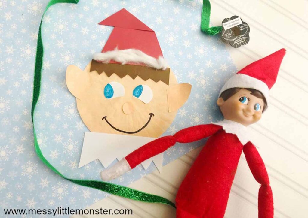 A crafted paper elf head with googly eyes and a red hat sits next to an elf doll in a red suit, perfect for Elf on the Shelf crafts. The backdrop is light blue adorned with white stars, and a green ribbon adds a festive touch.