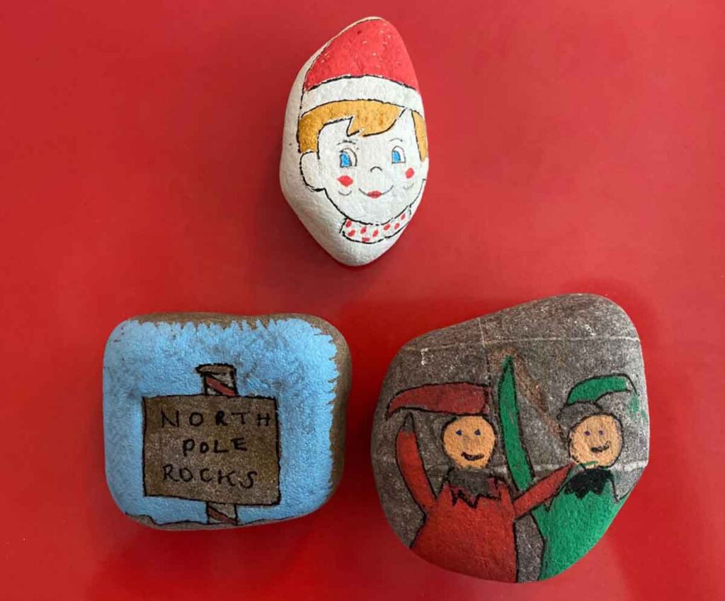 Three painted rocks rest on a red surface. The top rock features a smiling face with a red hat. The bottom left rock displays a "North Pole Rocks" sign, while the bottom right rock showcases two figures in festive red and green outfits, reminiscent of charming elf on the shelf crafts.
