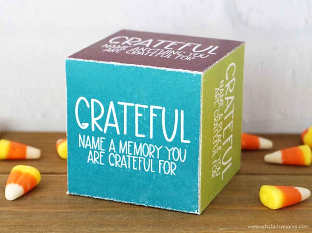 A cube with colorful sides displays the word "GRATEFUL" and prompts to name a memory to be grateful for. Candy corn pieces are scattered around the cube.