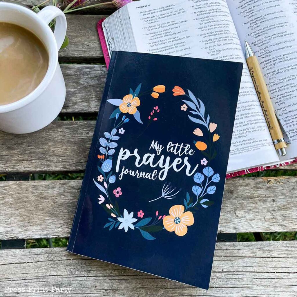 A floral-themed "My Little Prayer Journal for women" rests on a wooden table beside an open book and a pen. A cup of coffee is placed nearby, creating a cozy, reflective setting.