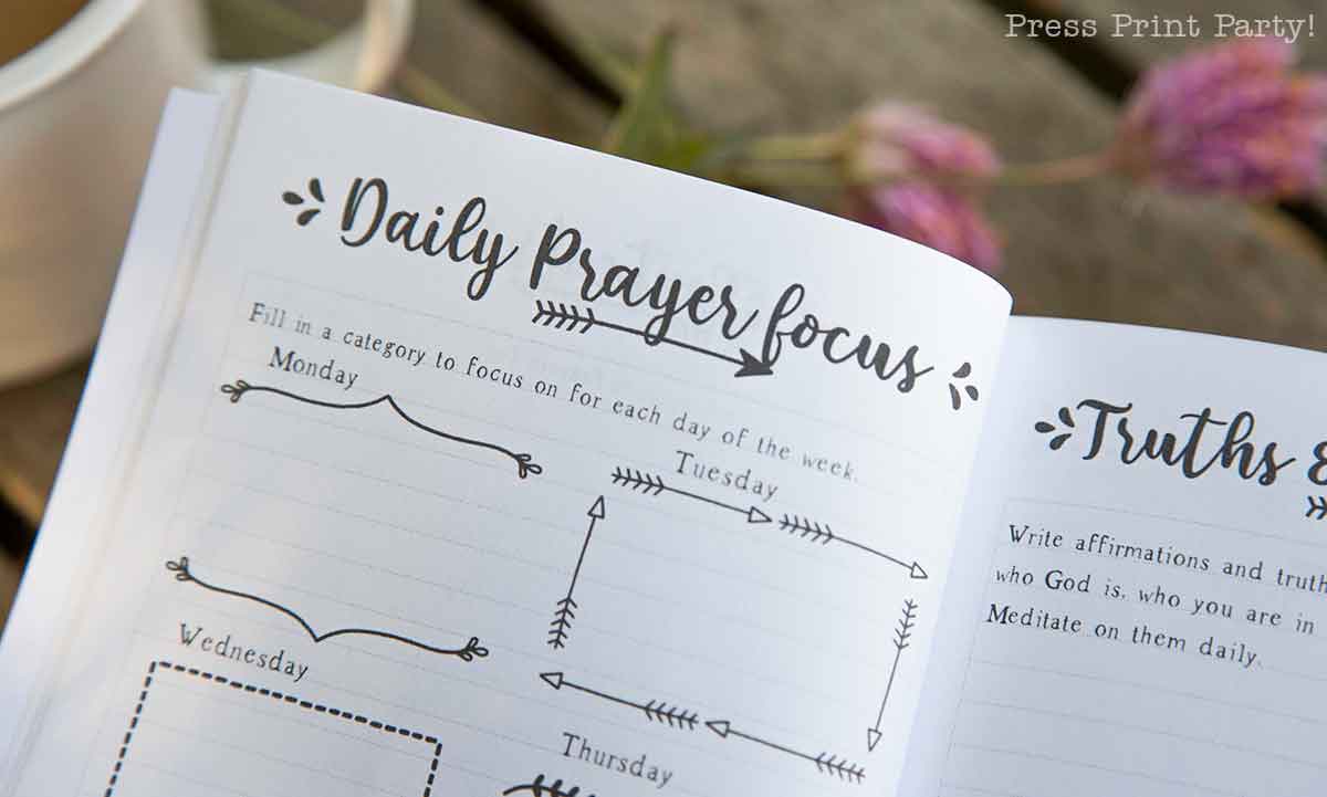 Open planner with a section titled "Daily Prayer Focus," resembling a prayer journal for women. It includes spaces for each day of the week to fill in categories. The page features decorative elements and rests on a wooden surface with a blurred plant in the background.