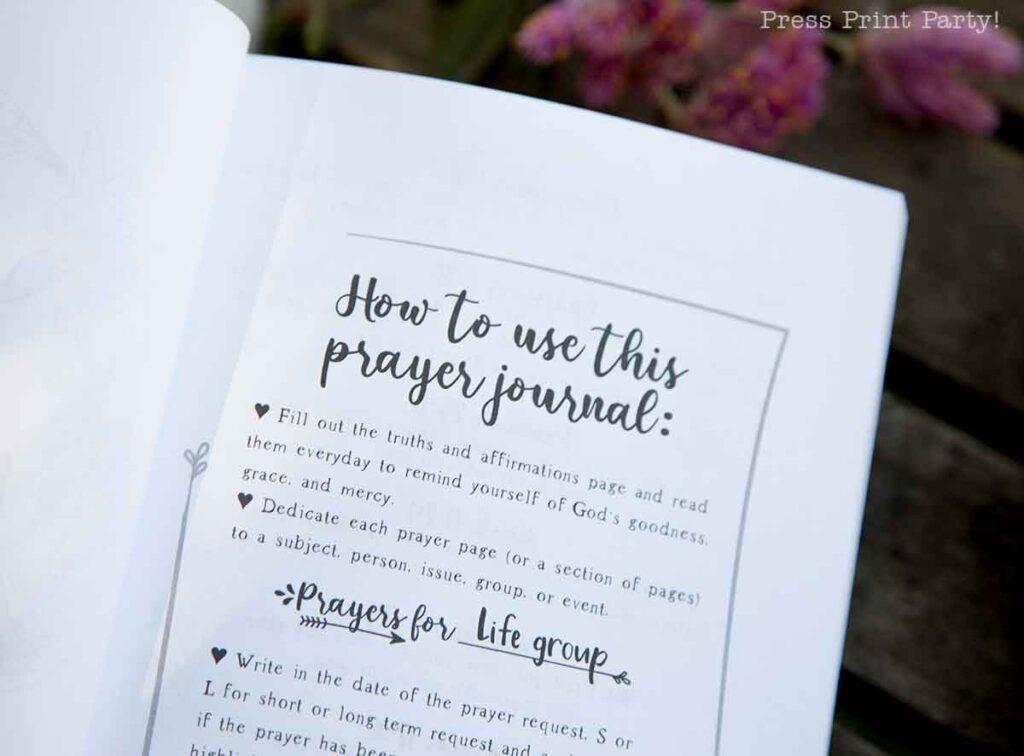 A close-up of an open prayer journal for women reveals instructions on its use, featuring sections for truths, affirmations, and dedicated prayer pages. The text sits gracefully against a wooden surface with purple flowers softly adorning the background.