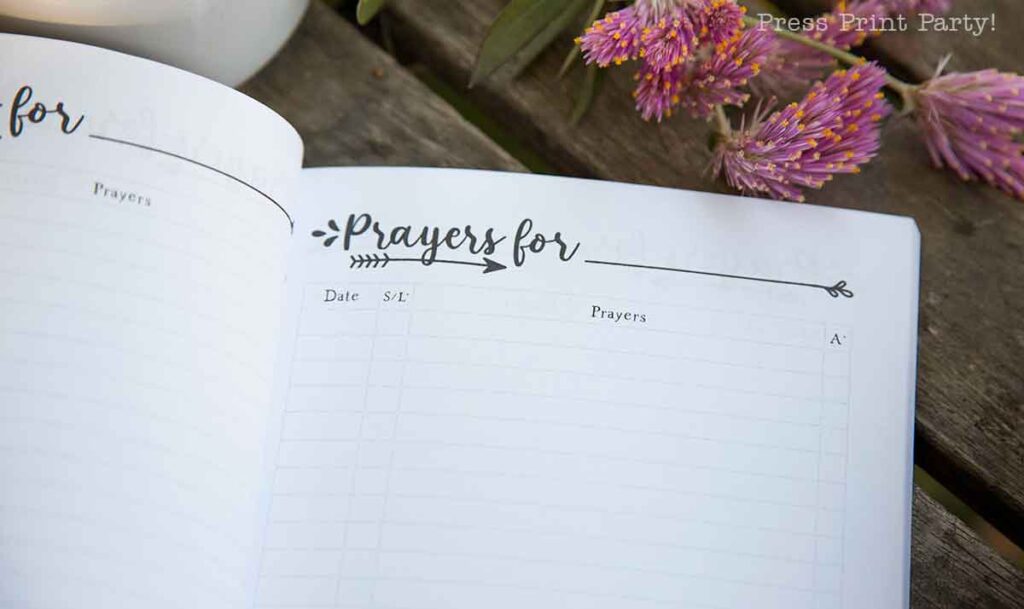 An open prayer journal for women rests on a wooden surface, its heading "Prayers for" inviting reflection. Neatly organized columns for "Date," "S/L," and "Prayers" await entries, while pink flowers sit gracefully beside it, adding a touch of serenity.