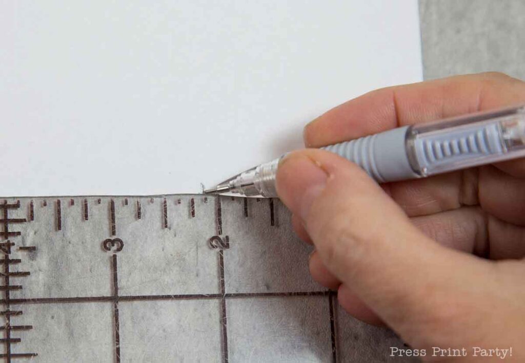 A hand uses a mechanical pencil to make a mark on a piece of white paper, with a pilgrim bonnet resting nearby. A clear ruler runs along the paper's edge, showcasing measurements in centimeters.