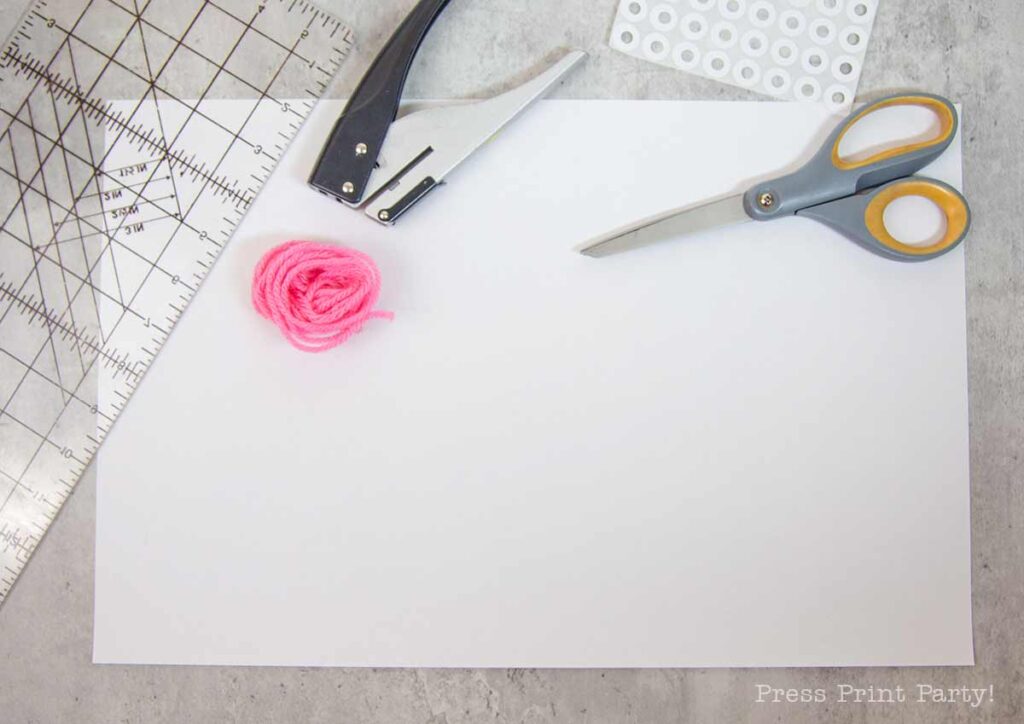 A crafting workspace with a sheet of white paper, a metal ruler, a pair of scissors, and adhesive reinforcement stickers set on a gray surface—a perfect setup for assembling projects like pilgrim bonnets using pink yarn and a trusty hole punch.