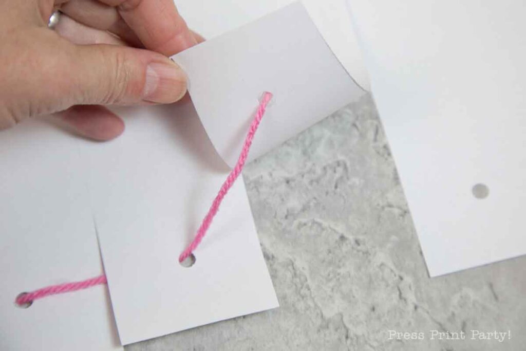 A hand delicately holds a piece of white paper, reminiscent of crafting a pilgrim bonnet, as pink yarn threads gracefully through a hole. The paper lifts slightly to reveal the intricate threading process against a serene light gray background.