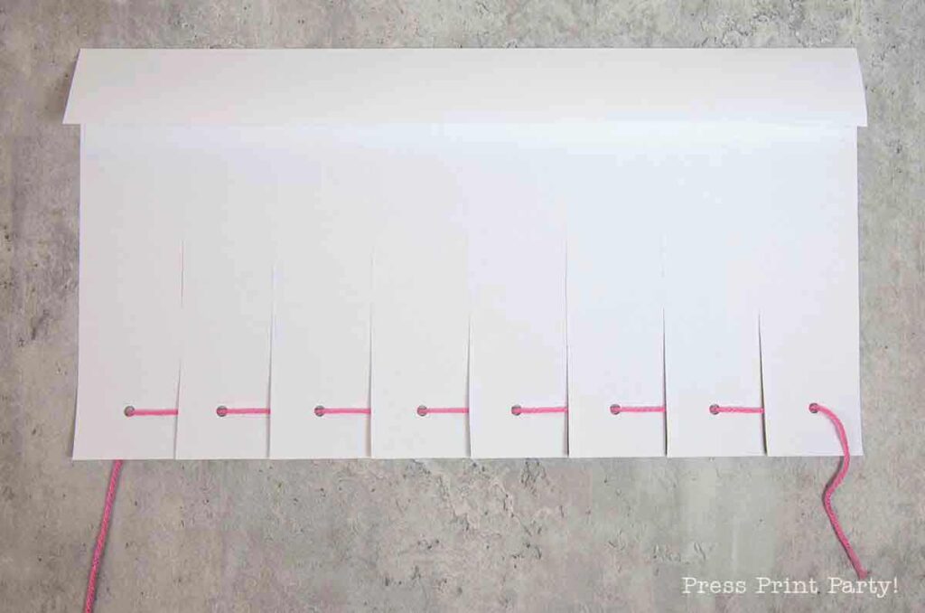 A white paper tent resembling a pilgrim bonnet features evenly spaced slits along the bottom edge, threaded with a pink string through nearby holes. A longer piece of string extends from one side against a textured light gray background.