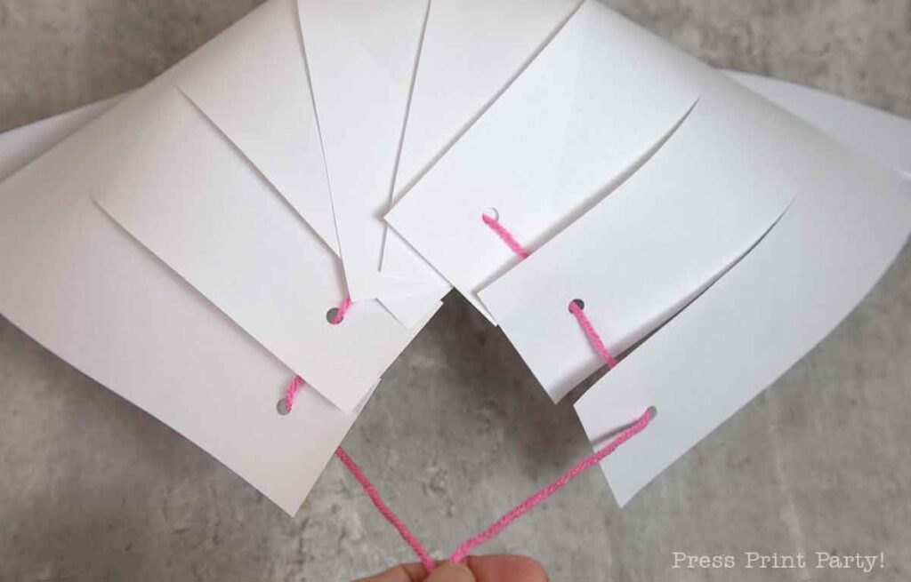 A handmade paper accordion craft features evenly spaced holes, threaded with pink yarn. Reminiscent of a pilgrim bonnet, the pages fold in a fan-like manner, and as the yarn is pulled, it elegantly gathers the paper together.