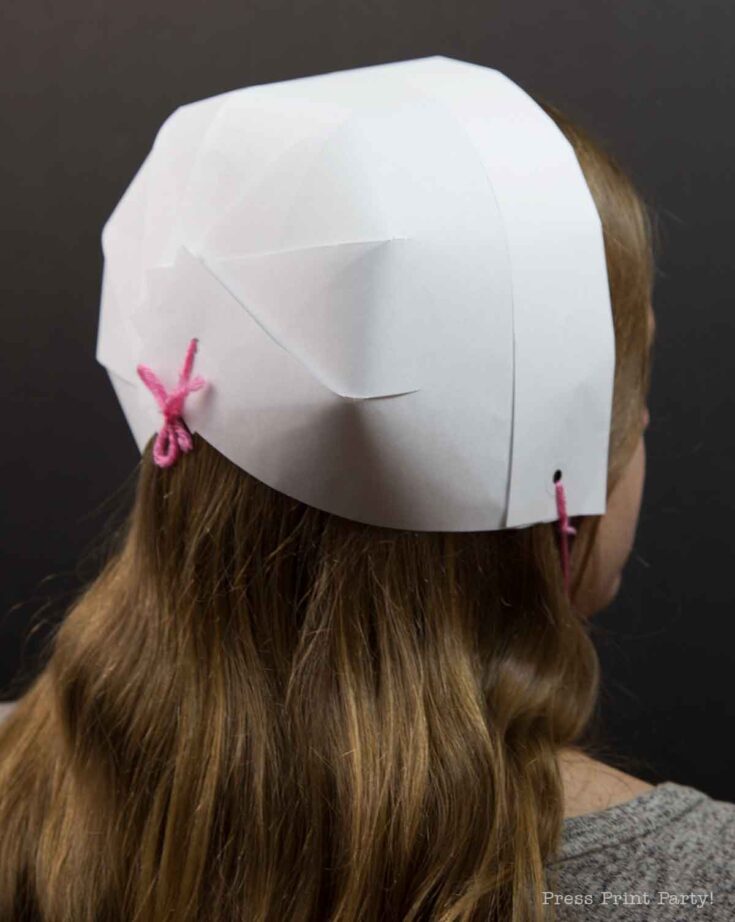 A person with long hair is wearing a pilgrim bonnet, crafted from white paper and secured with a pink string, shown from the back. The bonnet features overlapping sections and a neat bow at the nape, standing out against the plain dark background.
