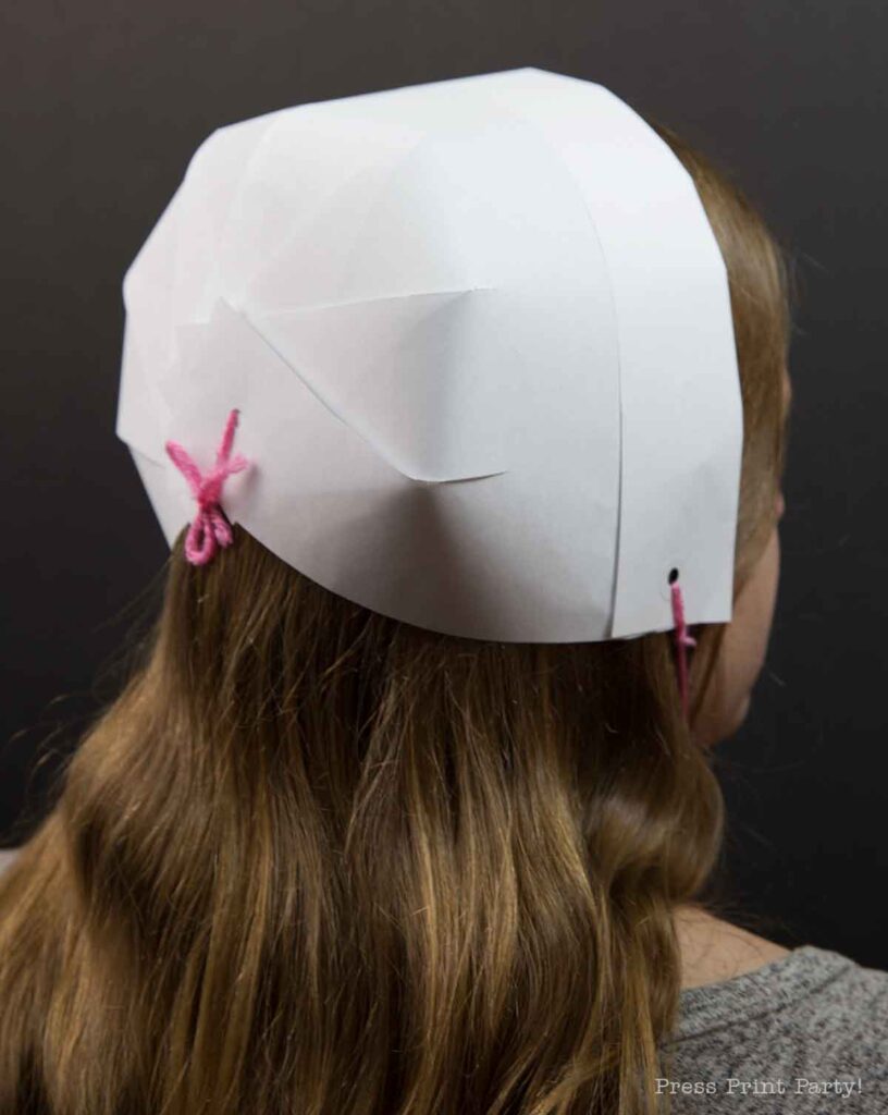 A person with long hair is wearing a pilgrim bonnet, crafted from white paper and secured with a pink string, shown from the back. The bonnet features overlapping sections and a neat bow at the nape, standing out against the plain dark background.